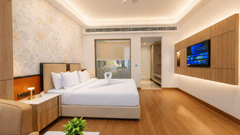 A spacious hotel bedroom featuring a double bed, a glass partition wall, a wall-mounted television, and wooden flooring at Pride Premier Greater Noida.