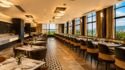 An indoor hotel restaurant containing arranged dining tables, cushioned chairs, and large glass windows showing trees outside at Pride Plaza Indore.