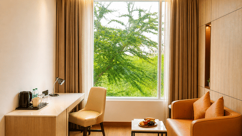 A room view at Pride Premier Greater Noida featuring a writing desk with a chair, a sofa, a small table, and a window looking out at trees.
