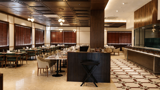 A dining hall at Mountnoor Sarovar Premiere, showing a buffet counter area, a tiled floor section with geometric patterns, and numerous dining arrangements. The space features large windows with horizontal blinds and a dark, structured ceiling.