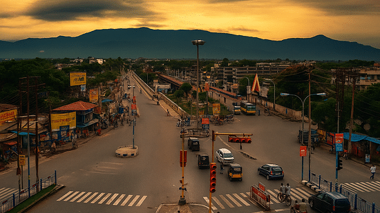 A busy city intersection in Siliguri, framed by distant hills, traffic, and pedestrians under a glowing sunset sky.