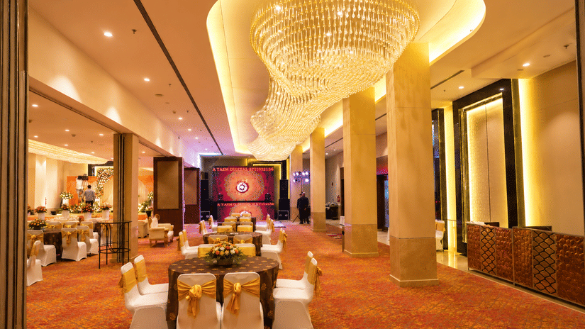 Spacious banquet hall with a chandelier ceiling, dining tables, and a stage area at Ambrosia Sarovar Portico, Haridwar.