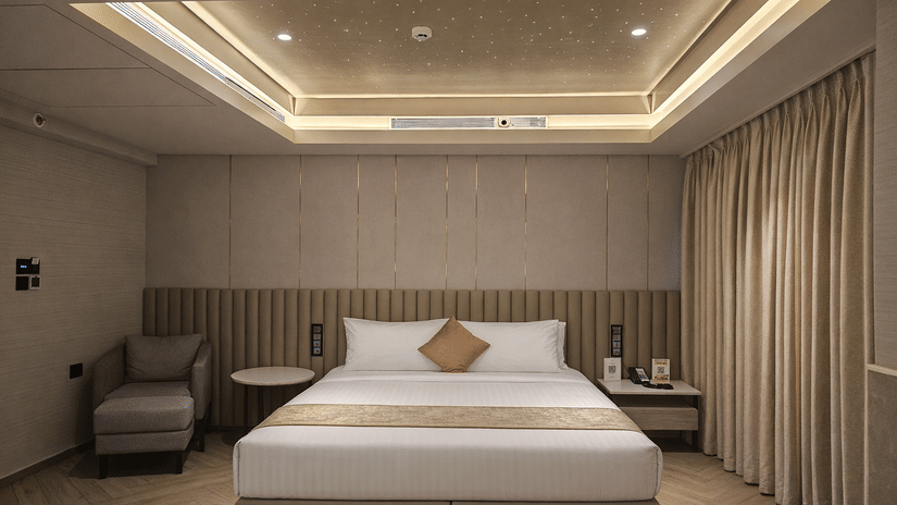 Double Seater Bathtub Room at Dragonfly Hotel – The Art Hotel with large bed, white linens, beige runner, accent pillow, nightstands with lamps, upholstered armchair, recessed tray ceiling lights, and beige curtains.