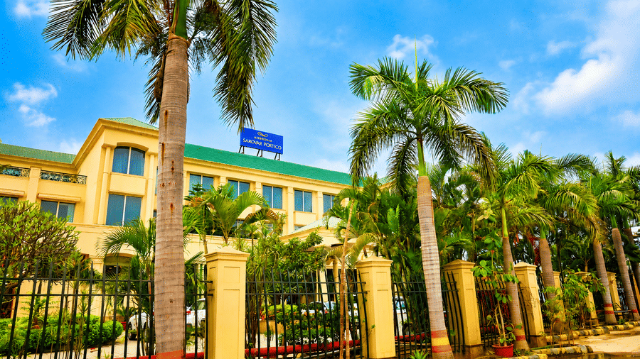 Front facade of Ambrosia Sarovar Portico, Haridwar, with a palm tress lined entrance and boundary gate view.