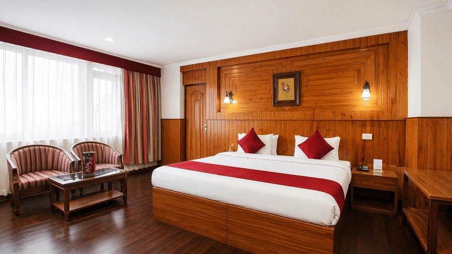 A room at Central Hotels featuring a king bed with red and white bedding wooden wall panels a cushioned armchair large windows with curtains and dark hardwood flooring.