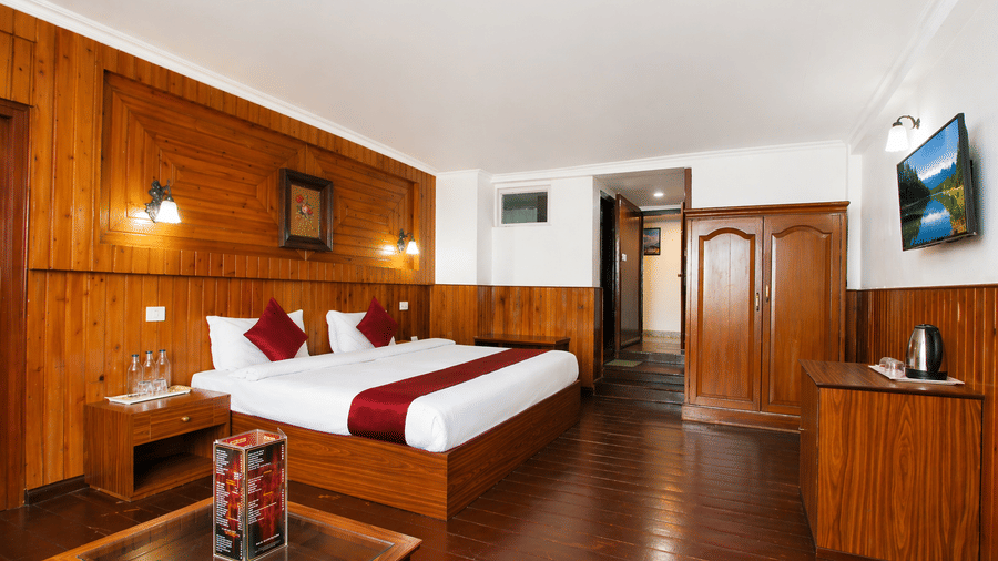 Wide angle view of a room at Central Hotels showing a king bed with red accents wooden wall panels dark hardwood flooring a work desk with kettle and wall-mounted TV near the door.