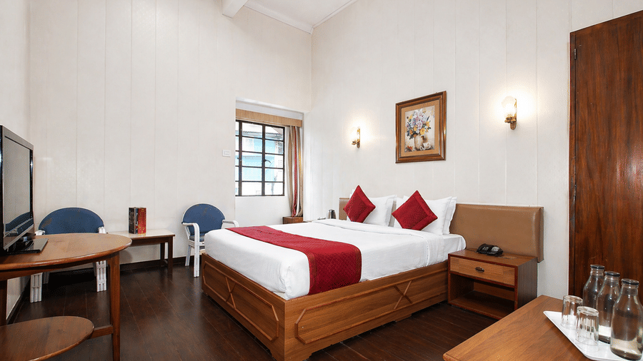 A spacious room at Central Hotels featuring a king bed with red and white bedding wooden furniture dark hardwood flooring a work desk with chair wall-mounted TV and large windows.