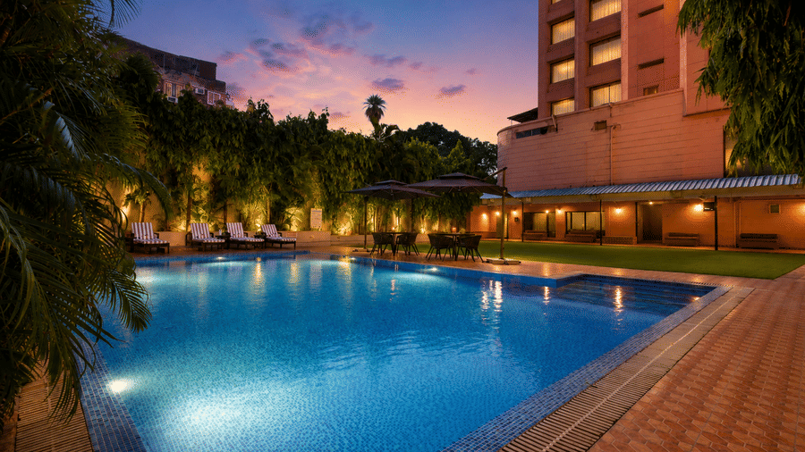Illuminated hotel pool at dusk with lounge chairs, tropical greenery, and warm ambient lighting.
