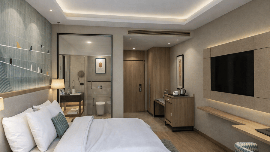 A modern bedroom at KPG Sarovar Portico, Raj Nagar Extension, Ghaziabad, showing a king size bed in the foreground and a wall-mounted flat-screen television opposite a wooden vanity desk.