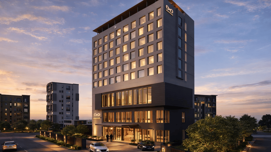 The KPG Sarovar Portico, Raj Nagar Extension, Ghaziabad, building at dusk, featuring many illuminated windows and 2 cars parked near the entrance under a sky with soft clouds.