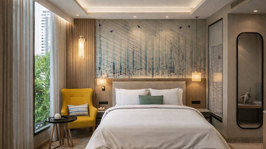 An interior view of a KPG Sarovar Portico, Raj Nagar Extension, Ghaziabad, hotel room showing a neatly made king size bed, a small side table, and a view of greenery through the window.
