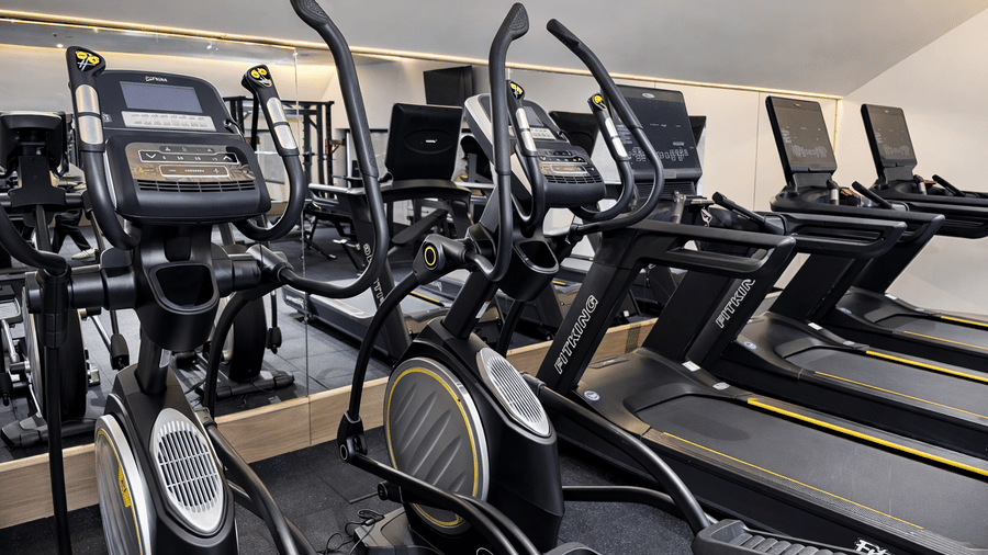 Gym at Mountnoor Sarovar Premiere featuring a row of treadmills and elliptical cross-trainers positioned in front of a full-length mirror, set in a room with sloped ceilings.