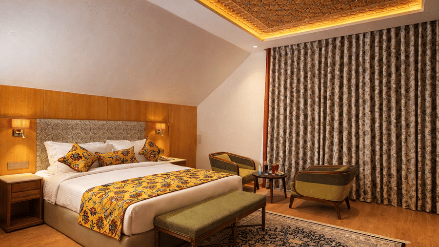 A Junior Suite at Mountnoor Sarovar Premiere with a king size bed, an ottoman at its foot, and 2 upholstered armchairs placed near a patterned rug.