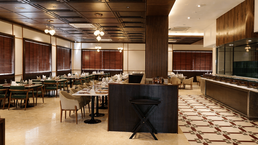 A dining hall at Mountnoor Sarovar Premiere, showing a buffet counter area, a tiled floor section with geometric patterns, and numerous dining arrangements. The space features large windows with horizontal blinds and a dark, structured ceiling.