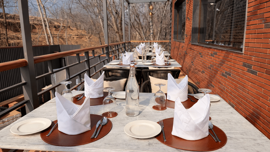 Outdoor seating area at Noor All-day Dining in Mountnoor Sarovar Premiere, Pahalgam, featuring set tables neatly arranged along safety railings offering views of the hills and the surroundings.
