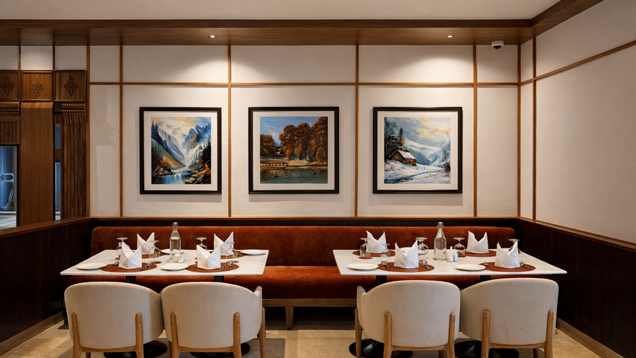 2 dining tables with 4 chairs placed in pairs on a side, and a sofa attached to the wall on which art pieces have been hung at Noor All-day Dining in Mountnoor Sarovar Premiere, Pahalgam.