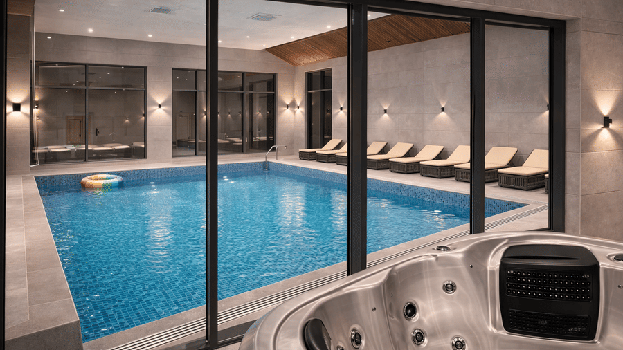 Indoor pool of Mountnoor Sarovar Premiere, Pahalgam, seen through paned glass windows, and lounge chairs placed on the pool deck.
