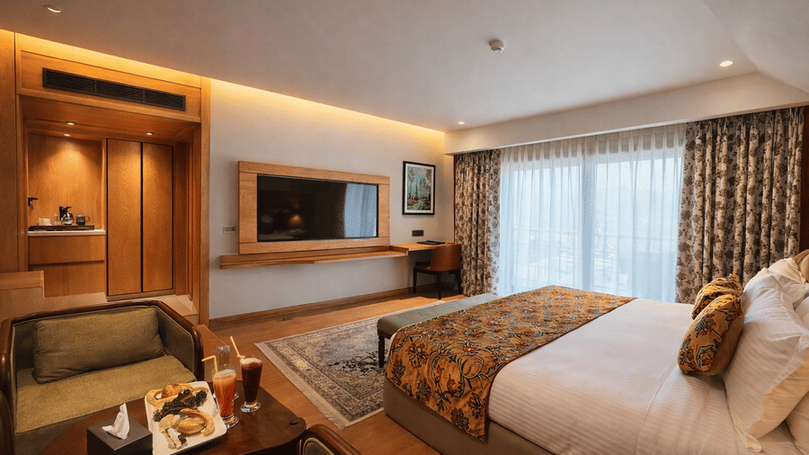 A view from the foot of a bed at Mountnoor Sarovar Premiere, showing a wall-mounted television and 1 small sofa with a tray of food on a table.
