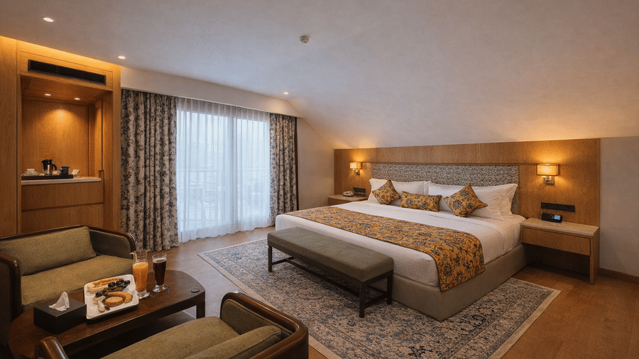 A spacious bedroom at Mountnoor Sarovar Premiere featuring a king size bed, a seating area with 2 chairs, and a large window with heavy drapes.