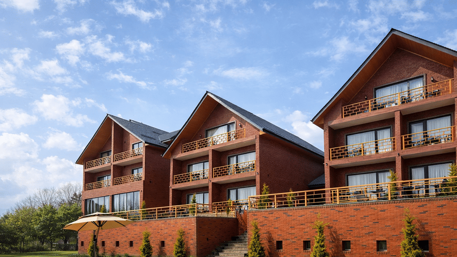 Multi-storey structure of Mountnoor Sarovar Premiere, Pahalgam, built of red bricks, with glass windows and tiled roof seen near a manicured lawn in which a table is set.