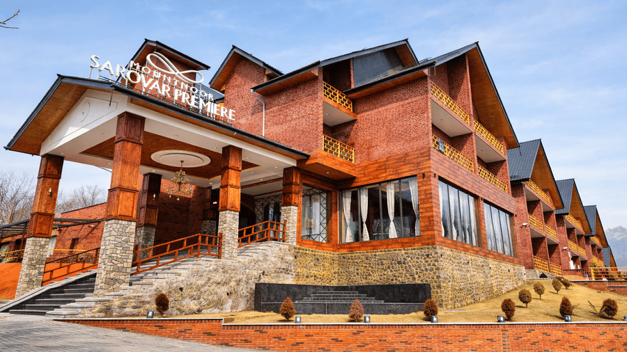 Lower angle of the entrance stairs and driveway of Mountnoor Sarovar Premiere, Pahalgam, showcasing its structure built by bricks, signage of the resort's name, and tiled roof.