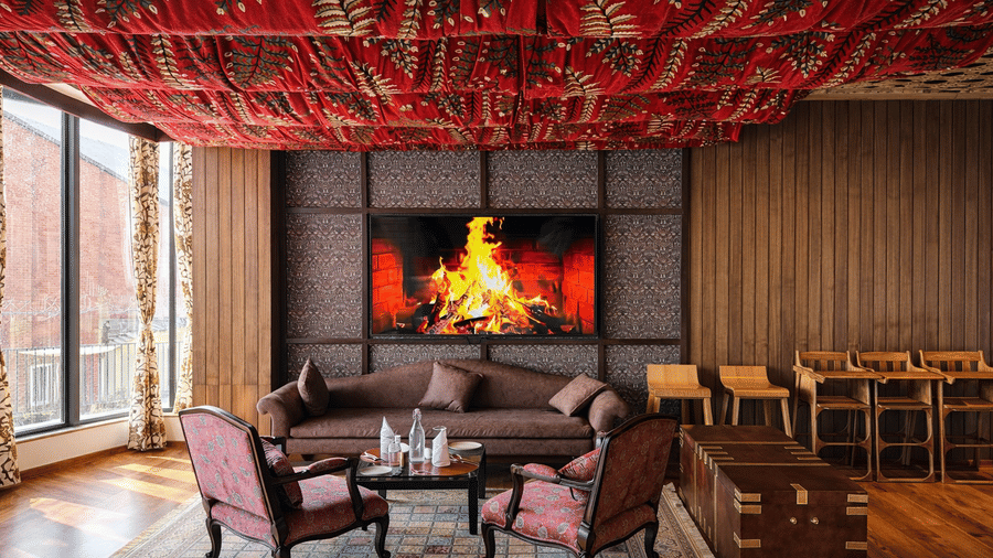 A lounge area in Tamanna Kashmiri Speciality Lounge at Mountnoor Sarovar Premiere, Pahalgam, with 2 armchairs facing a couch placed near a wall on much a television is displaying a lit bonfire.