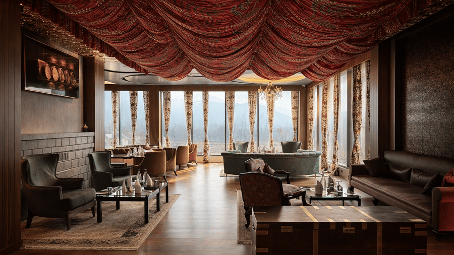 Wide view of the seating area and dining setup of Tamanna Kashmiri Speciality Lounge at Mountnoor Sarovar Premiere, featuring neatly arranged tables, armchairs, sofa, clothed ceiling, and large glass windows.