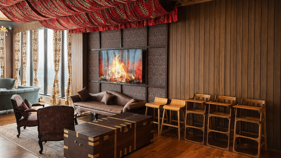 A dining space within Mountnoor Sarovar Premiere showing a mixture of plush sofas, traditional armchairs, and a trunk-style coffee table. A digital screen displays a fireplace scene beneath a draped fabric ceiling.