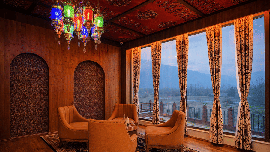 A seating table for 4 at Tamanna Kashmiri Speciality Lounge in Mountnoor Sarovar Premiere, placed near large glass windows offering views of the hills, and Turkish mosaic lamps hung overhead.
