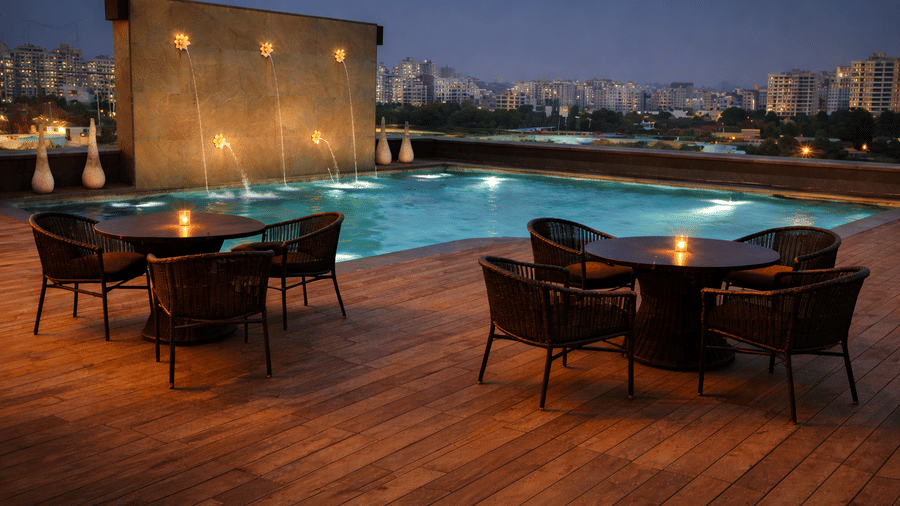 Illuminated rooftop pool deck at dusk featuring outdoor dining tables and chairs on wooden flooring.