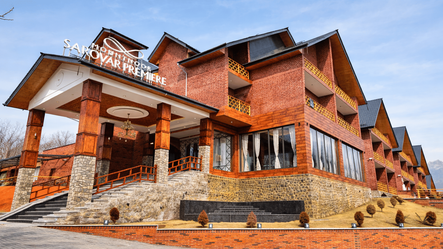 Lower angle of the entrance stairs and driveway of Mountnoor Sarovar Premiere, Pahalgam, showcasing its structure built by bricks, signage of the resort's name, and tiled roof.