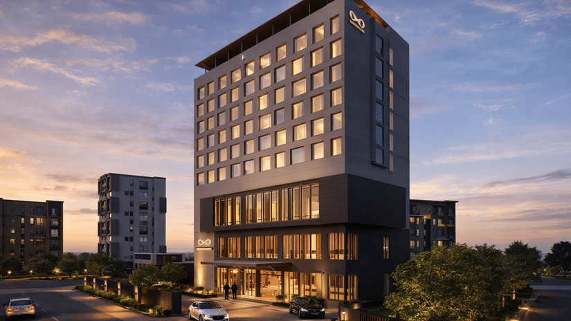 The KPG Sarovar Portico, Raj Nagar Extension, Ghaziabad, building at dusk, featuring many illuminated windows and 2 cars parked near the entrance under a sky with soft clouds.