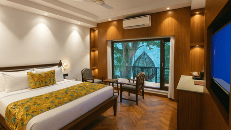 Premium Ganges View Room at Summit by the Ganges Beach Resort and Spa, Rishikesh featuring a double size bed, a sitting area with chairs and centre table, a cabinet table, a flat TV screen, an AC, and a balcony attached which has a chair placed.