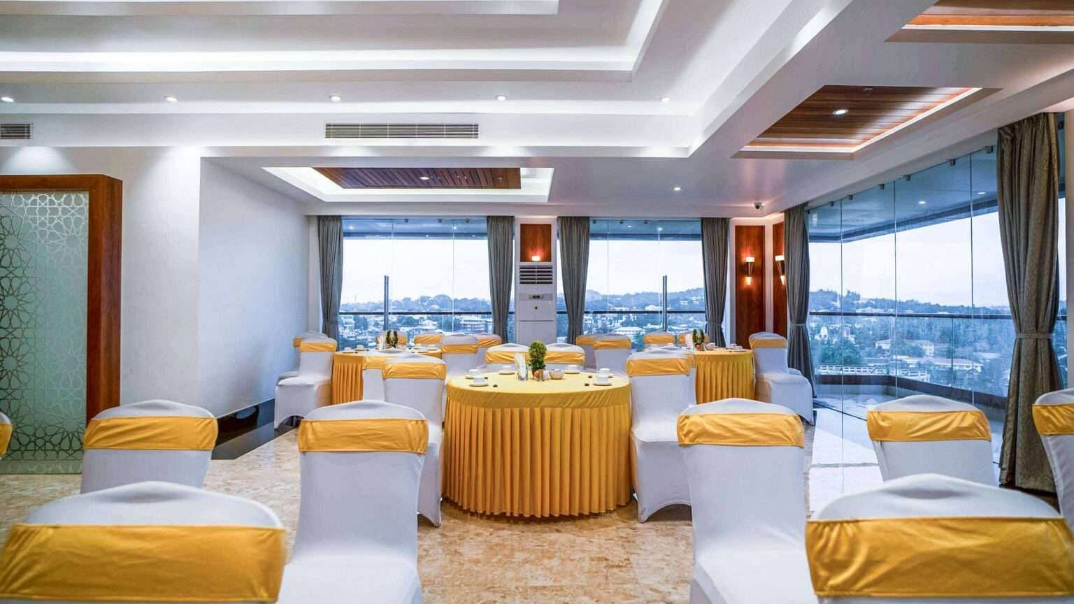 A brightly lit event space at Mansha Palace is set up with round tables and draped chairs, offering a city view.