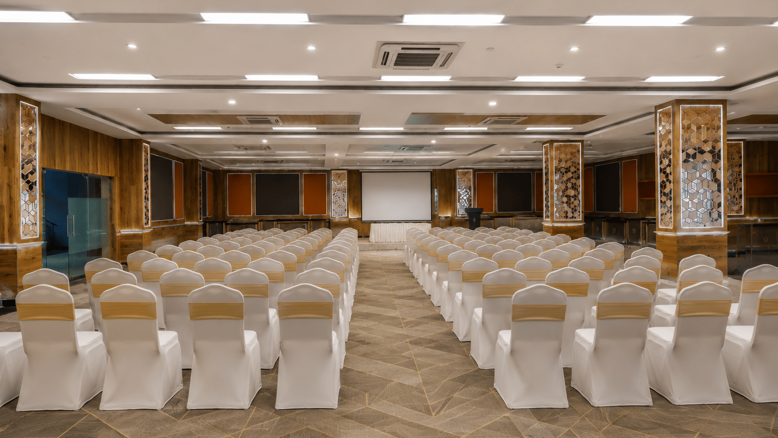 Celly banquet hall with theatre-style seating, white chair covers, a projection screen, podium and mirrored pillars at MAYFAIR World Cup Village, Rourkela.