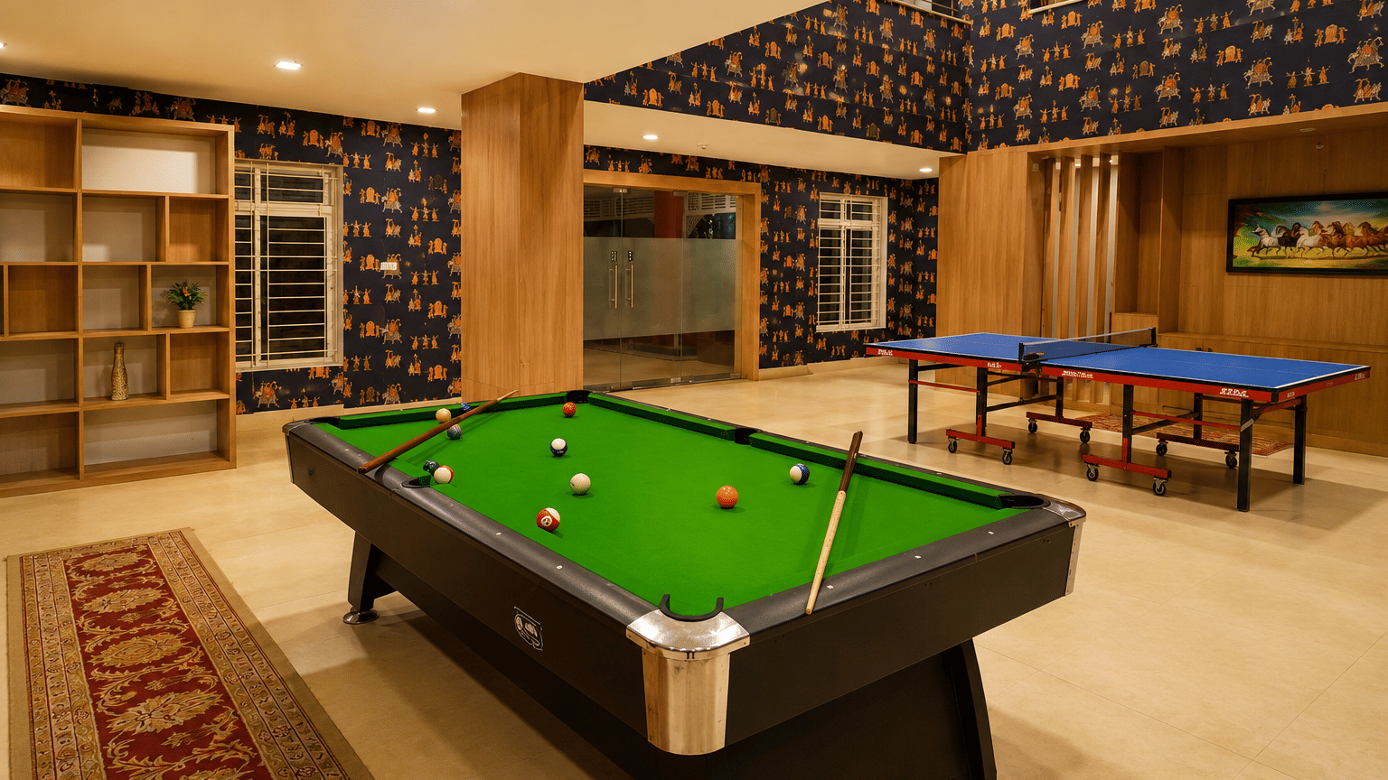 Indoor games room with a billiards table, two table tennis tables, wooden shelving and patterned wallpaper at MAYFAIR World Cup Village, Rourkela.