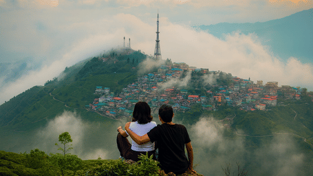 A couple sitting on a hilltop overlooking the misty mountains and colorful cityscape of Kurseong, with clouds floating over the valley, creating a premium and aesthetic morning vibe.
