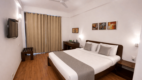 A bedroom with a king size bed, wall-mounted television, ceiling fan, and a large window at EMBLEM - Arjun Marg, Golf Course Road, Gurgaon.