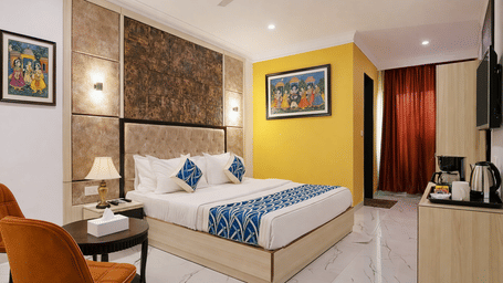 A deluxe room at Hotel Apple Park Delhi featuring an orange armchair, a double bed with a tufted headboard, and vibrant artwork on a yellow feature wall.