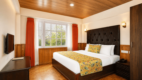 Deluxe Room at Orsino Khangri Karpo Retreat & Spa, Lachung, featuring a neatly-made bed, curtains framing a large window, wooden finishes and a television.