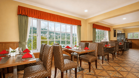 Orion restaurant at Orsino Khangri Karpo Retreat & Spa, Lachung featuring wooden tables and chairs with patterned upholstery stand on a tiled floor. Windows with curtains overlook the hills. Place settings include napkins and glassware.