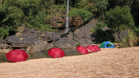 Riverside picnic area at Polo Resort Cherrapunji with row of red umbrellas and a blue tent on sandy bank by a waterfall.