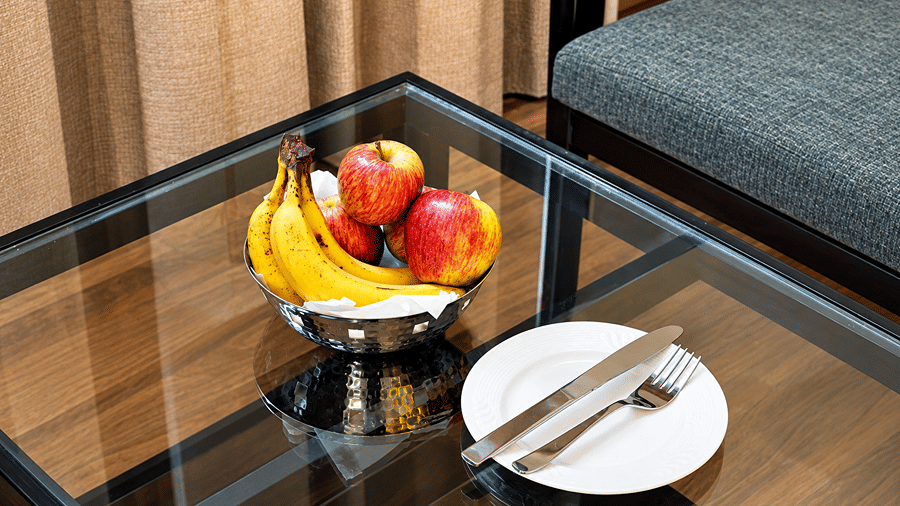 Fruit bowl with bananas and apples on a glass table beside a plate with a knife and fork at Daksh The Royal Braj, Vrindavan.