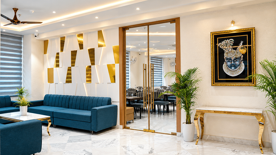 Lobby seating area at Daksh The Royal Braj, Vrindavan, featuring blue sofas, golden wall panels, a glass partition, and potted plants.