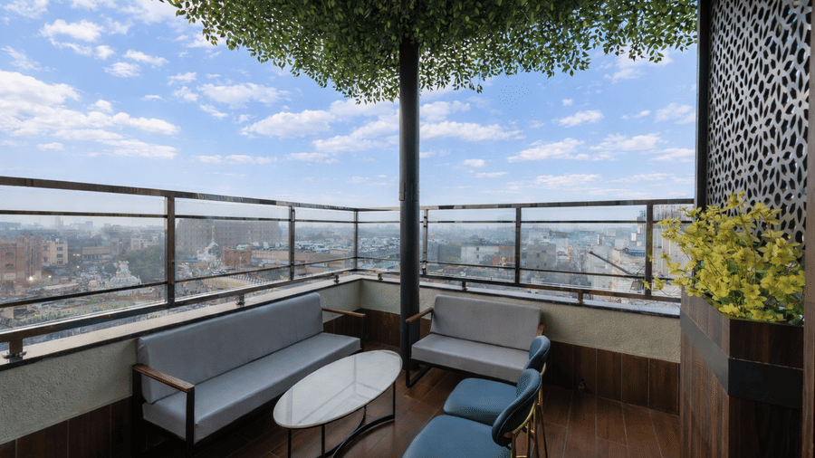 A balcony seating area at DLS Hotels Amritsar, with chairs, a sofa,  a table, potted plant, and a view of buildings and sky in the background.
