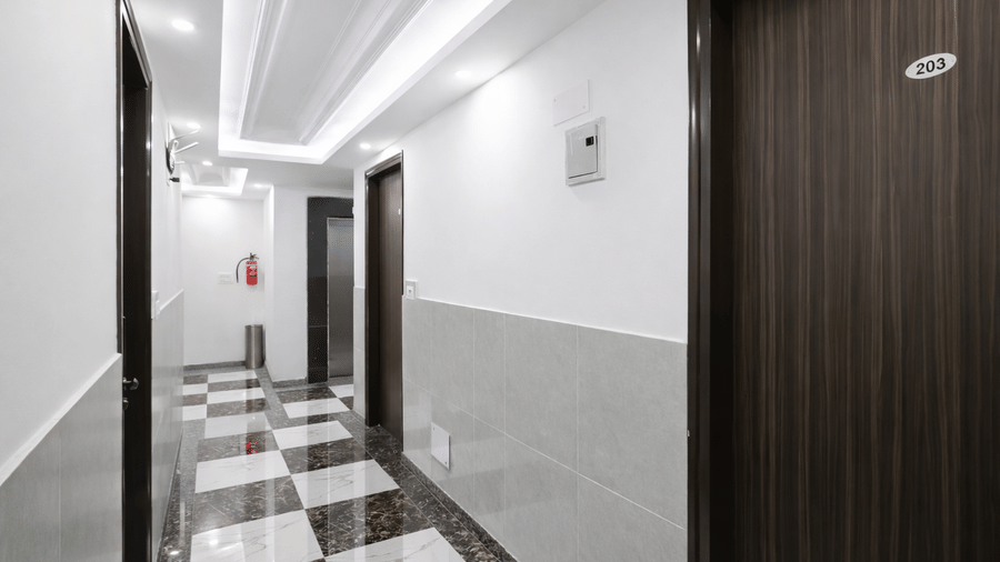 A brightly lit guest hallway at Hotel Apple Park Delhi with dark wood doors, grey lower wall panels, and polished chequered flooring.
