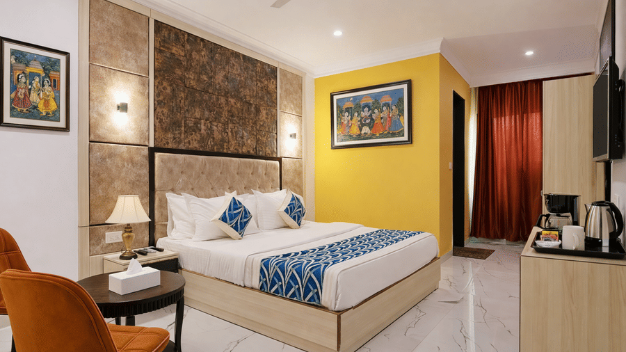 A deluxe room at Hotel Apple Park Delhi featuring an orange armchair, a double bed with a tufted headboard, and vibrant artwork on a yellow feature wall.