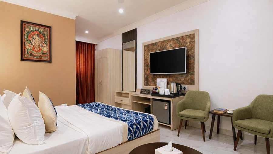 A guest room at Hotel Apple Park Delhi featuring 2 sage green upholstered chairs, a dark wood coffee table, and a television unit with a mini-bar.