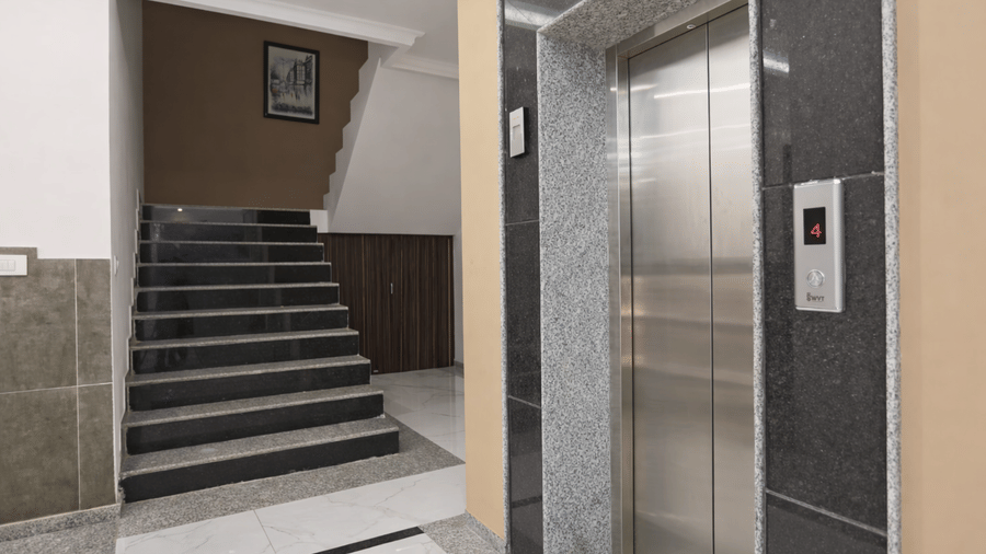 The lift area at Hotel Apple Park Delhi, showing stainless steel lift doors with a black marble surround and an adjacent flight of grey stone stairs.