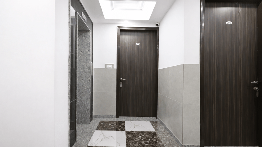 A hallway at Hotel Apple Park Delhi with two dark wood-finish guest room doors, white walls, and a polished chequered tile floor.
